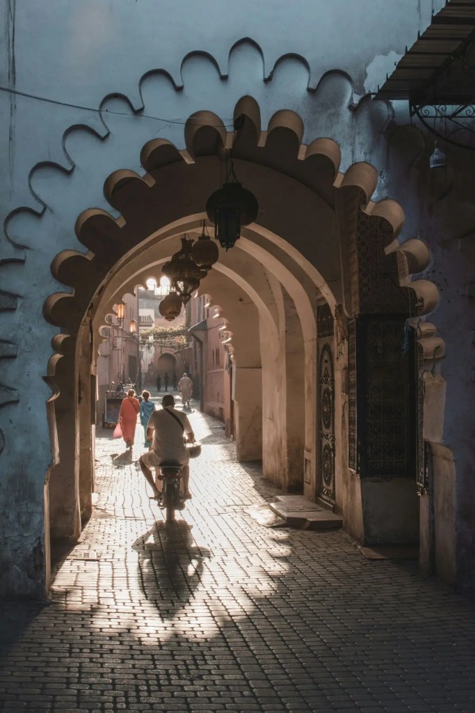 People walking through a shaded, narrow tunnel alleyway in the Marrakech Medina.