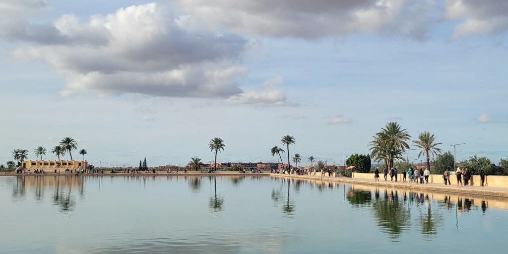 A panoramic view across the large reflecting basin at the Menara Gardens in Marrakech. The still, greenish water acts as a massive mirror, perfectly reflecting the partly cloudy blue sky, several tall palm trees, and the small figures of people walking along the distant, sunlit promenade.